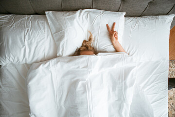 Young teen girl under blanket in white bed in bedroom with gesture V sign. Healthy sleep, sleep apps, lazy morning