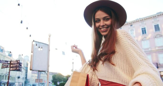 Portrait of beautiful Caucasian girl in hat looking at camera and smiling outdoor. Joyful nice Caucasian young woman in good mood standing on street. Happiness and positive emotions. Leisure concept