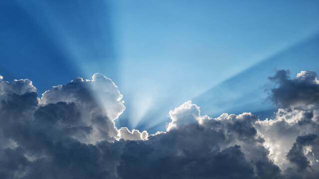 Sun rays behind dark clouds in a blue sky, horizontally panoramic
