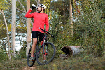 Cyclist resting and drinking isotonic drink in tropical forest, wears bike helmet and red cycling...