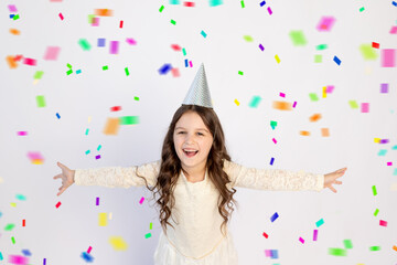 A cute little girl with a cap on a white isolated background with a streamer is happy with her arms outstretched. Space for text. Little girl celebrates birthday, holiday concept