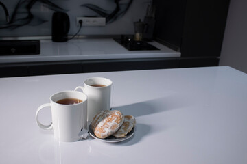 two white cups with tea and sticking out tea bags next to gingerbread cookies heart-shaped on a white table in a gray kitchen. High quality photo