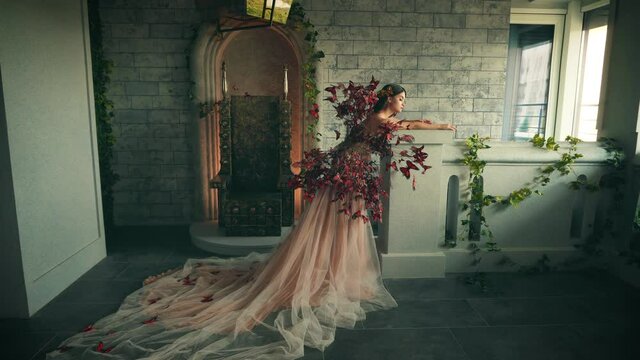 Beautiful young fantasy woman in image magic fairy princess. Elven Queen stands in ancient medieval room. Creative costume red butterfly. beige peach luxury fairytale dress, long lush train plume