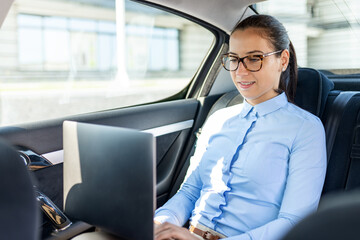 Young business woman in the back seat of the car, typing on laptop.