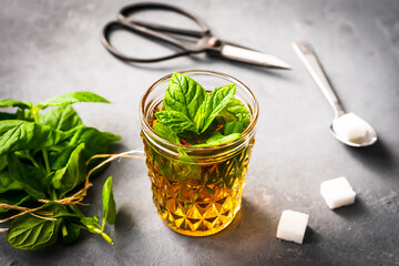 Pfefferminze in einem Glas, Würfelzucker, Löffel und ein Bund frische Minze auf einem grauen Küchentisch. Teepause, rustikaler Stil.