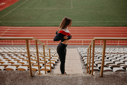 Smiling girl stands at the city stadium, lifts up a sports jacket, and shows her muscles. Mativation concept. Lady with six pack. Progress result after diet and exercise. Image with copy space.