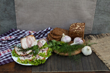 On a white plate on lettuce leaves are bacon with pepper, dill and parsley. Black bread, garlic and herbs lie next to it on a round board..