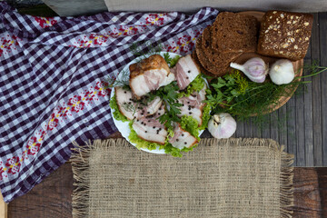 View from above. Against the background of a checkered napkin on the lettuce, there is bacon with pepper, dill and parsley. Nearby, on a round board, lies black bread, garlic and herbs.
