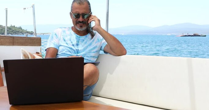 Portrait of a freelancer businessman is sitting on the deck of a yacht with a laptop computer and smartphone at summer vacation