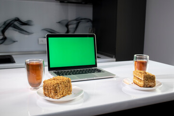 two cups of tea and two pieces of cake in gray kitchen next to laptop with green screen - chromakey. High quality photo