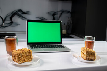 two cups of tea and two pieces of cake in gray kitchen next to laptop with green screen -...