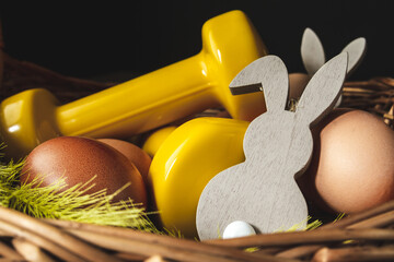 Traditional Easter wicker basket with eggs, decorative bunny and yellow dumbbells. Easter fitness and training composition.