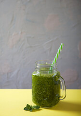 A green detox smoothie in a glass jar with a straw stands on a yellow table on a gray background. Healthy eating.