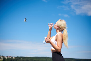 Young blond fit woman, wearing black leggings and white top,drinking water from glass bottle outside with blue sunny sky on background. Female fitness training in summer. Healthy lifestyle concept.