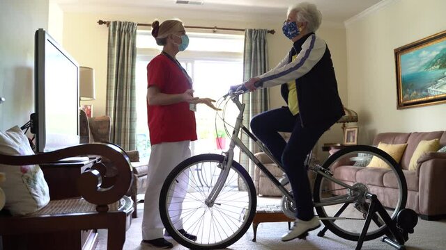 Low angle view of home visit nurse working with elderly woman on exercise bicycle in front of television, both wearing face masks.