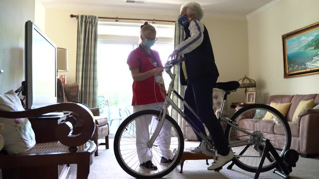 Senior elderly woman gets onto exercise bicycle and home visit nurse helps her in a living room in front of television.