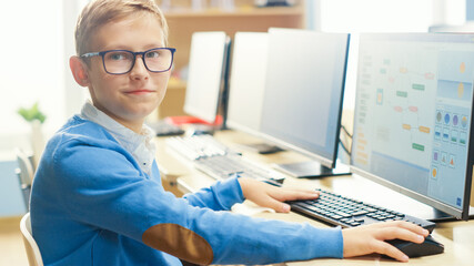 Elementary School Computer Science Classroom: Cute Little Boy wearing Glasses Uses Personal Computer, Learning Programming Language for Software Coding, Turned and Looks at Camera.