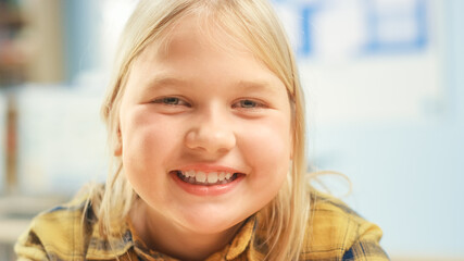 Portrait of a Cute Little Girl with Blond Hair Sitting at her School Desk, Smiles Happily. Smart...
