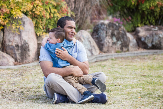 Aboriginal father sitting on grass hugging mixed race aboriginal caucasian son