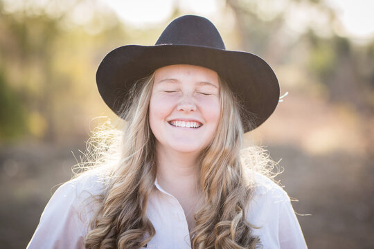 Teenage girl laughing with eyes closed wearing akubra hat on farm in drought