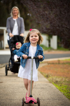 Little girl on scooter on footpath with mother behind