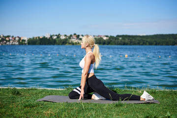 Young blond woman, wearing black leggings and white top, doing fitness stretching exercises on grey yoga mat by lake in summer morning. Female sport training outside on fresh air. Healthy lifestyle.