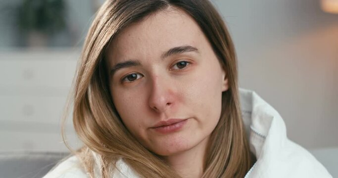 Close up of ill young Caucasian woman looking at camera and blowing nose in napkin while sitting at isolation at home. Portrait of unhealthy girl with flu or coronavirus.