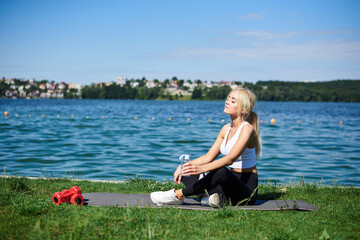 Young blond woman, wearing black leggings and white top, sitting on grey yoga mat by city lake in summer morning, relaxing resting after sport training outside on fresh air. Healthy lifestyle.