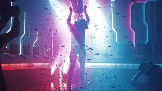 Professional Video Games Player Tournament Winner Celebrates Victory Cheering and Holding Trophy. Big Stylish Neon Bright Championship Arena doing Pro Computer Gaming Event with Online Streaming