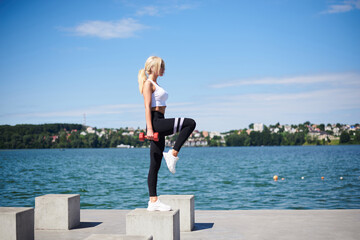 Fototapeta na wymiar Young blond fit woman, wearing black leggings and white top, training outside by city lake on concrete platform, holding red weights. Female power training in summer. Healthy lifestyle concept.