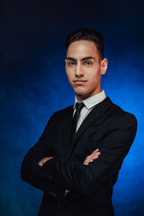 Handsome young man in elegant suit keeping arms crossed and looking on the camera. Studio shot on a dark background illuminated by blue light