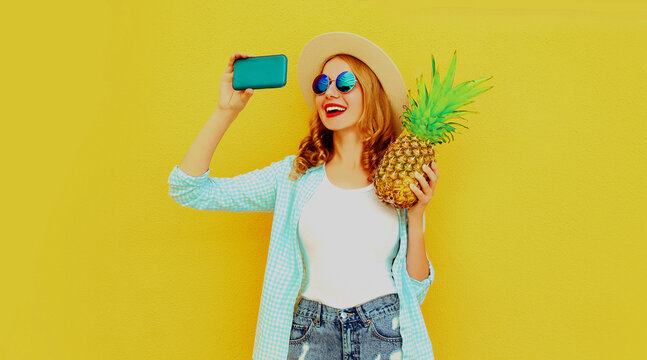 Summer portrait of happy smiling woman with pineapple taking selfie picture by smartphone wearing a straw hat, sunglasses over colorful yellow background