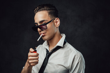 Handsome and successful. Young handsome man in sunglasses wearing white shirt and black tie lights up a cigarette and looking on the camera against black background