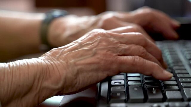 Closeup of elderly woman’s hands with arthritis typing on computer keyboard inside in bright sunlit room.