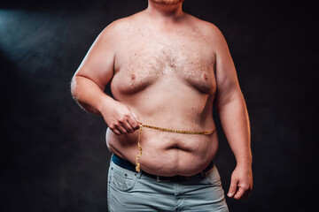 Chunky man is measuring his fat belly with a yellow measuring tape, standing on the black backgrund
