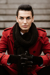 Fototapeta na wymiar LGBTQ community lifestyle concept. Young homosexual man sits on a stairs. Handsome fashionable gay male model poses in cityscape outdoors. Wears red coat, gloves, and black scarf