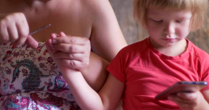 Young mother cuts nails of little blond boy. Child is busy playing on smartphone