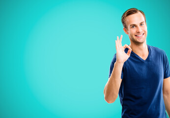 Portrait image of happy smiling man in blue casual clothing, showing okay gesture, or zero hand sign, aqua marine color background. Brunette excited male model at studio.