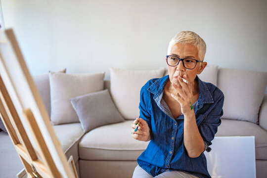 Beautiful mature woman painting and drawing in her home- art studio, while smoking a cigarette. Creative senior woman. Elderly woman is painting in her home. Retirement hobby.