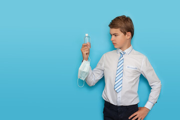 A teenage boy in a shirt and tie holds a bottle of sanitizer and a medical mask. Protecting health during training.