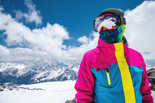 Portrait of a girl skier in a multi-colored bright jacket in a ski mask with her face closed on a sunny day against the backdrop of snow-capped Caucasian mountains and clouds
