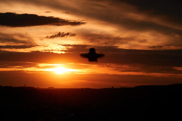 Small drone also multicopter as a silhouette out of focus. Orange evening sky with clouds, city in the background. Germany.