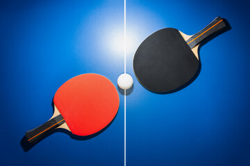 Top view black and red table tennis racket and a white ping pong ball on the blue ping pong table with a bright spotlight, Two table tennis paddle is a sports competition equipment for indoor exercise