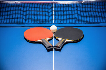 Black and red table tennis racket and a white ball on the blue ping pong table with a net, Two table tennis paddle is a sports competition equipment indoor activity and exercise for background concept