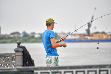 A young fisherman in a cap, blue T-shirt and shorts throws a fishing rod on the Volga river...
