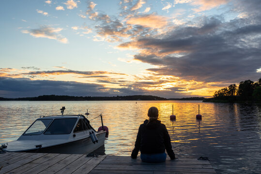 One woman sits with her back on the wooden pier at sunset and looks into the distance. Summer beautiful Baltic sea evening landscape. Horizon line with setting sun. Lonely Woman  on the coast near yac