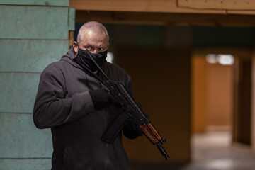 A man airsoft gunman in a black mask on his face posing with an akm machine gun in his hands in an...