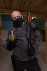 A man airsoft gunman in a black mask on his face posing with an akm machine gun in his hands in an...