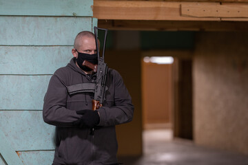 A man airsoft gunman in a black mask on his face posing with an akm machine gun in his hands in an...
