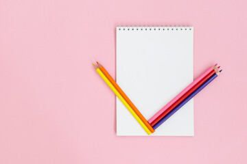 Multi-colored pencils on a white notebook. Composition on a light pink background, copy space.